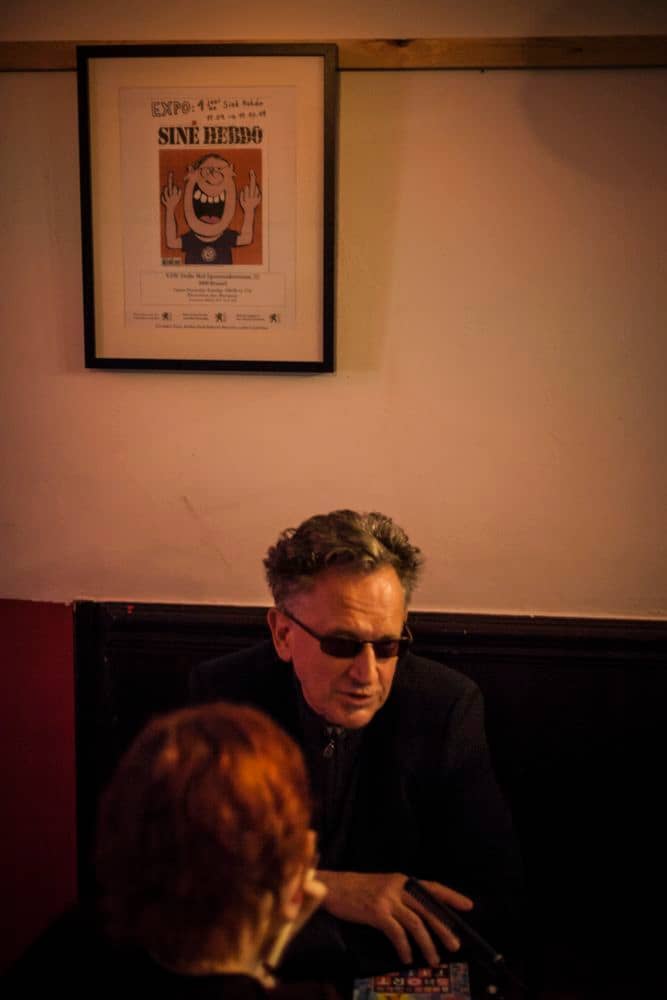 Dans un bar, Benoît Delépine parle. Derrière lui, la couverture du premier numéro du Siné Hebdo, encadrée.